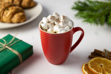 Red mug with hot drink and marshmallows. Green gift box, Christmas tree and croissants on the plate. White background
