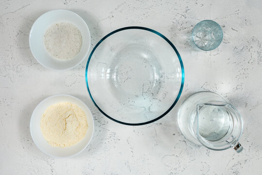 Glass and ceramic bowls with wheat flour, a glass and a jug of water for kneading dough on a white table.