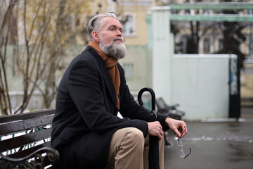 An adult gray-haired respectable man with glasses sits on a park bench and enjoys a winter morning