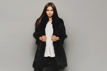 Young model woman with a bright manicure in an expensive winter fur coat posing on the grey background. Studio shot, winter fashion