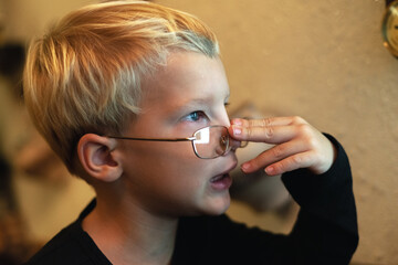 The preschool boy pulls his glasses over his nose with his finger and looks at something carefully. Close-up portrait
