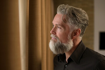 stylish adult bearded gray-haired man looks out the window, watching what is happening