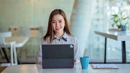 Young asian businesswoman beautiful charming smiling and using digital tablet while sitting at table and working in office. Looking at camera.