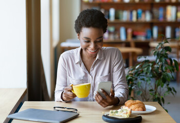 Fototapeta na wymiar Young black woman using smartphone for online communication while having lunch at coffee shop