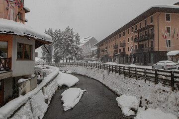 Small river Torrente Frigidolfo in the village of Santa Caterina in heavy snowfall. Visible hotels and houses with some snow flakes falling down.