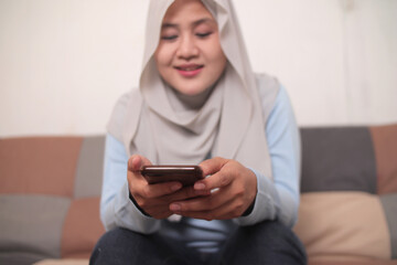 Asian muslim woman sitting of the sofa at home using phone and smiling. Female doing communication on her smart phone, sending message or browsing online