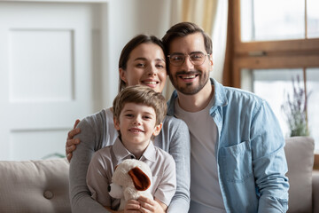 Portrait of happy young family with little son hugging plush teddy bear in own living room. Smiling parents mom and dad with small boy child relax on couch at home, show unity and bonding.