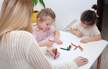 A young kindergarten teacher draws with children. Mom and daughters draw with pencils at the table.