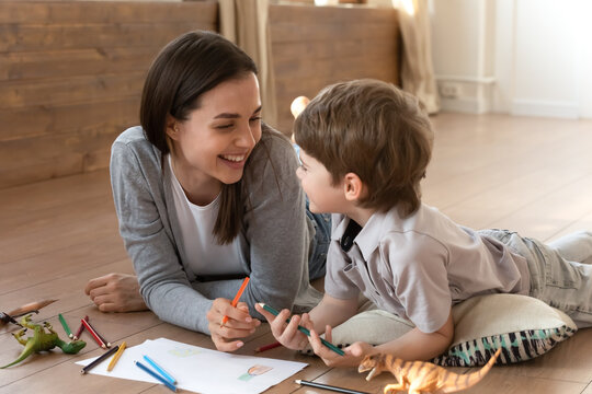 Happy loving young Caucasian mom and small son lying on floor in living room drawing painting together. Smiling caring mother or nanny have fun playing with little boy child on family weekend at home.