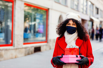 Obraz premium Portrait of a woman in red shopping outside on the street with face mask walking and holding many christmas gifts and presents