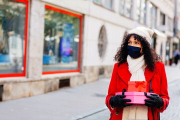 Obraz premium Portrait of a woman in red shopping outside with face mask holding many christmas gifts and presents looking at shop windows