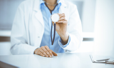 Unknown woman-doctor in blue blouse is holding stethoscope head while sitting at the desk in clinic, close-up. Physician ready to examine and help patient. Medicine concept