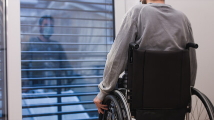 Fototapeta na wymiar Disabled lonely man wearing face mask sitting in a wheelchair alone looking through window.