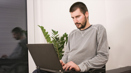 Young man in wheelchair with laptop working remotely from home, using internet and looking at...