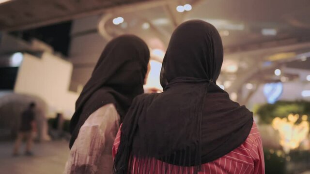 Two asian muslim friends standing in front of shopping mall, tourist pointing at map sign, window shopping customers consumerism, holiday travel vacation, big public community mall people walking pass