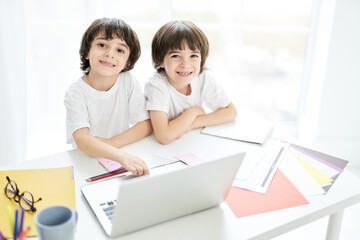 Best education for kids. Two adorable latin boys, brothers smiling at camera while sitting together at the table and using laptop. Little kids having online lesson at home