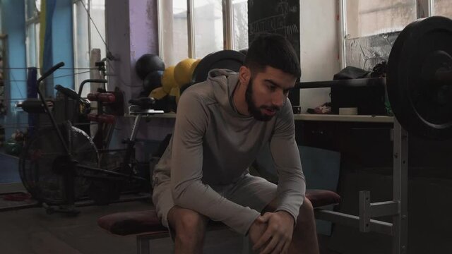 Sad, tired, weak, thin, little guy after a workout wipes sweat from his forehead with a towel in the gym. Barbell swing. Overall plan. Static camera