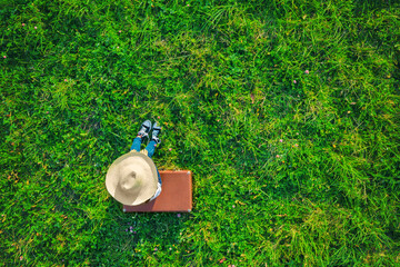 Aerial drone view of tired girl with straw hat and jeans sitting and resting on brown vintage suitcase on green grass meadow. Travel concept.