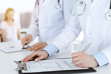 Fototapeta premium Unknown woman-doctors at work with patient at the background. Female physicians filling up medical documents or prescription while standing in sunny hospital reception desk, close-up. Health care