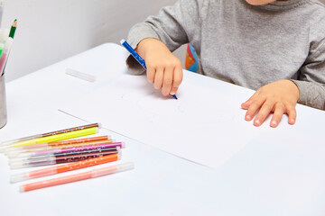 Cute little boy with blond hair draws colored pencils at home. Draws at the white table. Close up of child's hands drawing at white paper