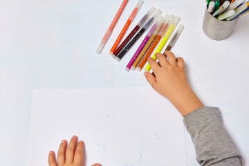 Cute little boy with blond hair draws colored pencils at home. Draws at the white table. Close up of child's hands drawing at white paper