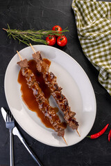 grilled beef skewers in meat sauce on a white plate on a dark background. Decorated with vegetables, herbs and textiles