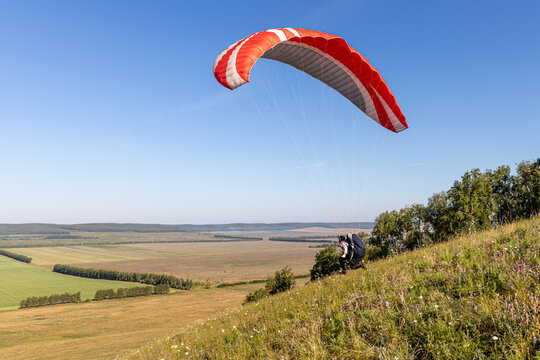Beautiful view of a pilot with red and white paraplane going to start flying over green and yellow fields in summer in the morning