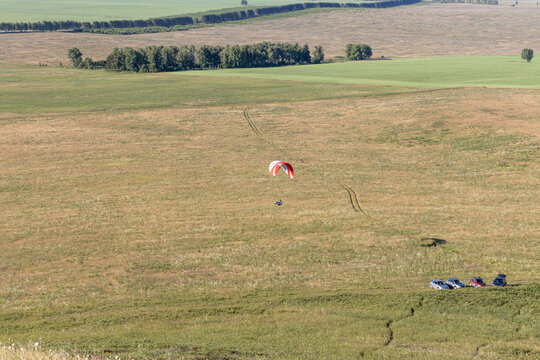 Beautiful view of a pilot with red and white paraplane flaying over green and yellow fields in summer in the morning