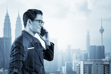 Fototapeta premium Prosperous handsome entrepreneur in suit and glasses pensively talking phone and looking on Kuala Lumpur cityscape. The concept of problem solving consulting . KL skyscrapers. Double exposure.