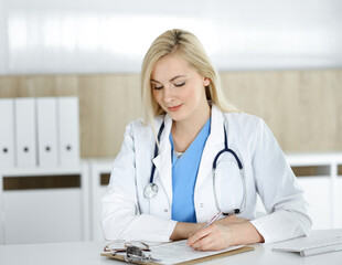 Woman-doctor at work while sitting at the desk in hospital or clinic. Blonde cheerful physician filling up medication history record form