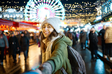 Obraz premium smiling woman at everning festive Christmas fair holding someone hand, follow me. ferris wheel on background. Happy winter holiday