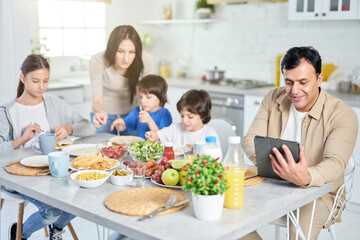 Obraz na płótnie Canvas Family routine. Smiling hispanic father using digital tablet while having lunch together with his family, sitting at the kitchen table at home
