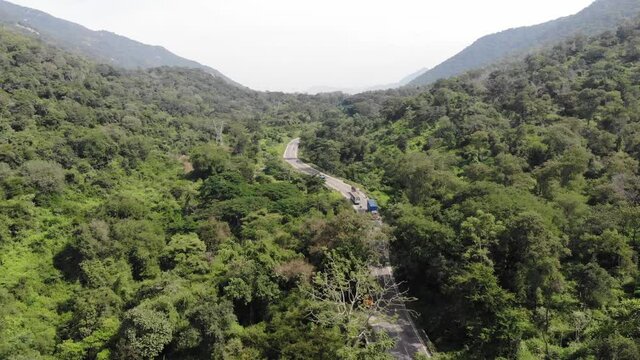 Aerial flyover indian asphalt highway with traffic surrounded by green nature forest mountains during sunny day.