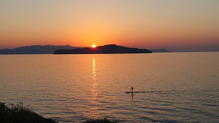 Naklejka premium A man is sailing on a board with an oar during a beautiful golden sunset over the water on Crete, Greece.