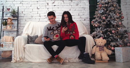 Fototapeta na wymiar Wide view. Young couple at home near the Christmas tree, using their mobile phone to track the New Year discounts. Bright living room with brick wall. Happy winter holidays