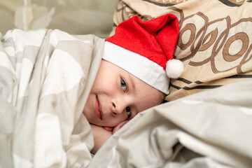 A satisfied, smiling boy lies in bed in a Santa hat. The joyful feeling of the holidays - New Year, Christmas.