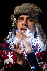 Fototapeta premium A young guy holds a garland in his hands. Christmas garland. The guy is holding a garland in front of him, a Christmas decoration. The light of the New Year's lights.