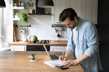 Serious man wearing glasses calculating domestic bills or taxes, household expenses, managing budget, paperwork, checking financial documents, using laptop and calculator in modern kitchen