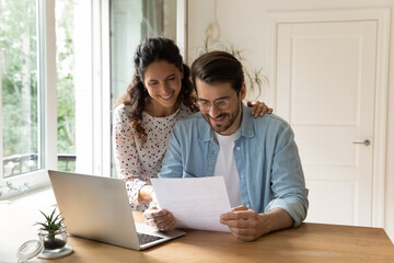Happy wife and husband wearing glasses reading good news in letter, sitting at table with laptop,...