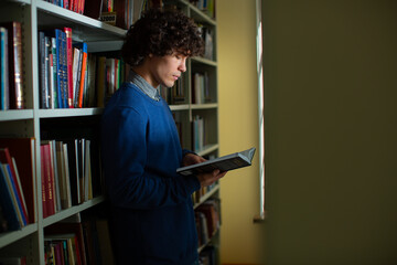 A focused fellow student with curly hair in a shirt, and blue sweater stands leaning on a bookshelf and reads a book in the library