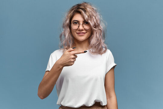Isolated image of beautiful stylish young woman in trendy glasses and white t-shirt posing against blank copy space wall background, smiling, pointing fore finger sideways. Body language and gestures