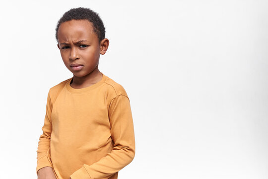 Angry dark skinned little boy in yellow sweatshirt frowning eyebrows, being stubborn. Displeased African child posing isolated against white studio wall background with copy space for your information