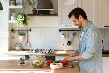 Side view smiling man cutting fresh vegetables, cooking homemade salad, standing at wooden table countertop in modern kitchen at home, preparing food for dinner or party, enjoying leisure time