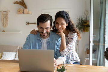 Happy young man wearing glasses and woman hugging, using laptop together, looking at screen, enjoying leisure time, spouses surfing internet, shopping online, making purchases, booking tickets