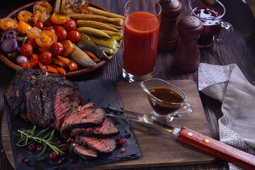 Appetizing roast beef with sauce and spices on a wooden table next to grilled vegetables and tomato juice