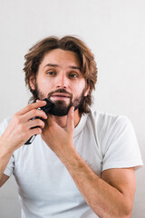 Handsome man shaving his beard with electric shaver in front of the mirror.