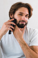Man shaving his beard in the mirror with electric razor. Man with beard. Shaving his beard in the...