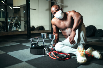 Fototapeta na wymiar Muscular african american male sitting on the floor in the gym in a medical mask with dumbbells, kettlebells and a training expander. Pandemic training
