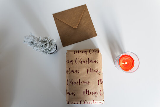 Close-up photo of a gift between a small Christmas tree and a candle with an envelope on top of a white table. During winter, thousands of gifts and Christmas cards are sent by post around the world.