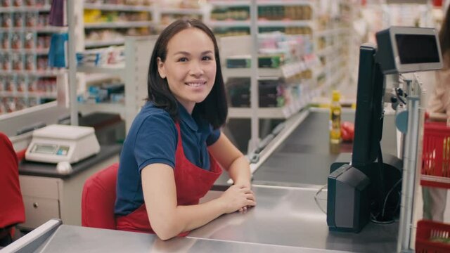 Medium portrait of friendly woman in uniform working as cashier in big supermarket smiling at camera while unrecognizable customer in mask placing products on conveyor belt in blurred background